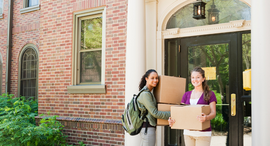 Two college students smiling and carrying boxes while moving into a dormitory, representing the excitement of starting college life.