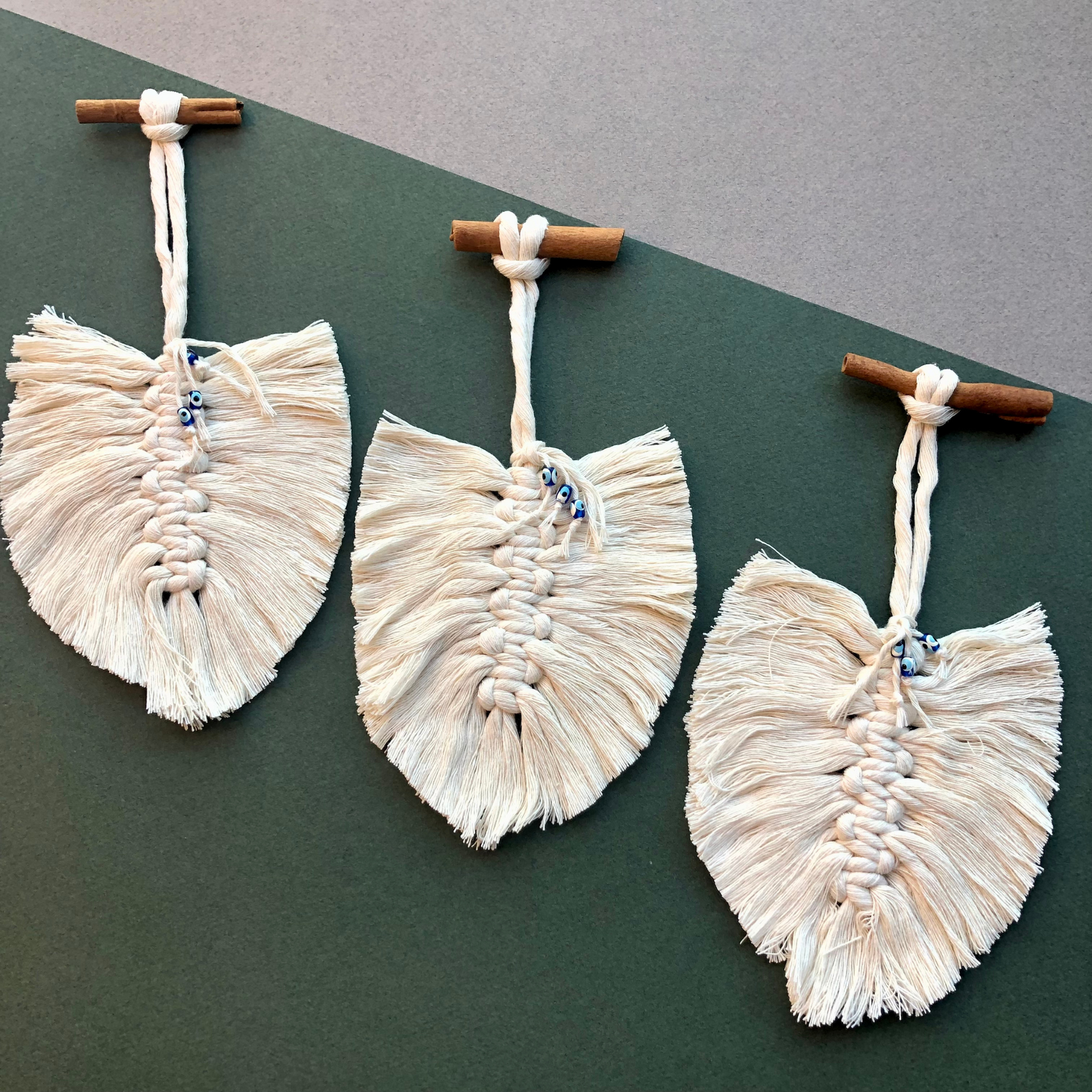 Set of 3 cream macrame feathers with cinnamon stick hangers on a dark green & grey background, ideal for adding a natural, bohemian touch to your decor