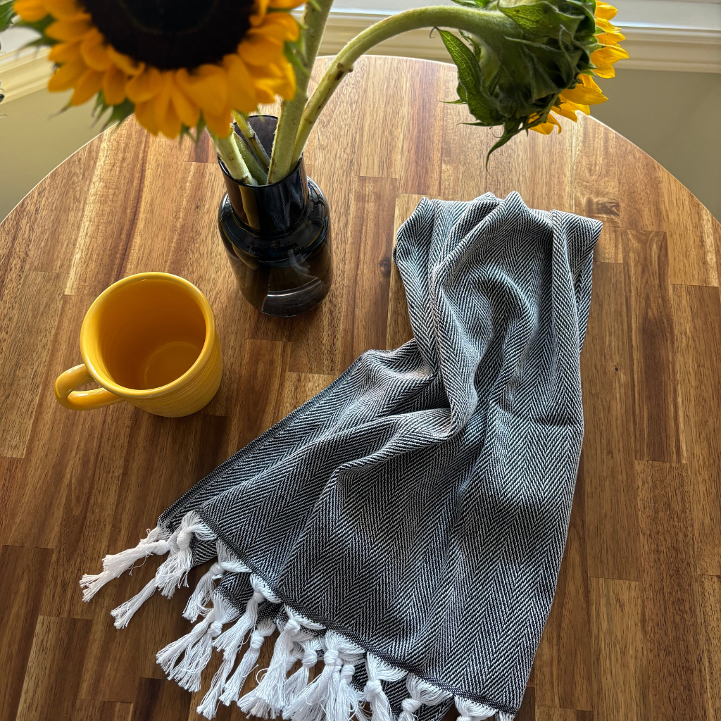 HERRINGBONE Turkish Kitchen Towel with white tassels draped casually on a wooden table next to a yellow mug and a sunflower in a black vase, creating a cozy kitchen setting