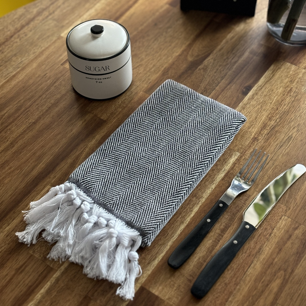 Neatly folded HERRINGBONE Turkish Kitchen Towel with white tassels placed on a wooden table next to a sugar jar, fork, and knife, offering a stylish dining setup