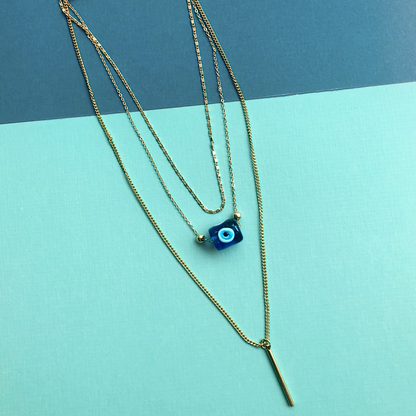 Layered evil eye necklace with gold chains, featuring a blue nazar charm and bar pendant on a turquoise and dark greenbackground