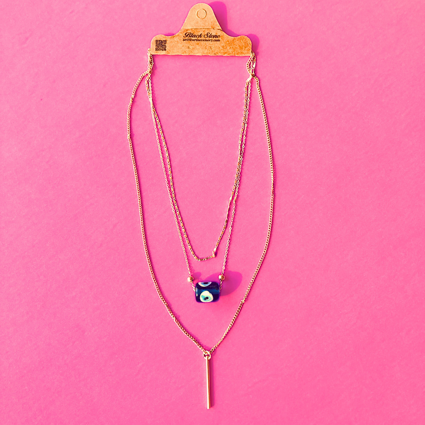 Layered evil eye necklace with a blue nazar charm and bar pendant displayed on packaging against a pink background