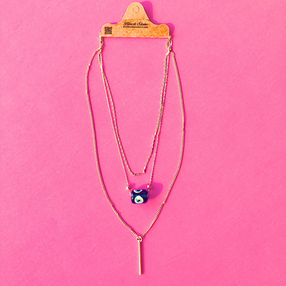 Layered evil eye necklace with a blue nazar charm and bar pendant displayed on packaging against a pink background