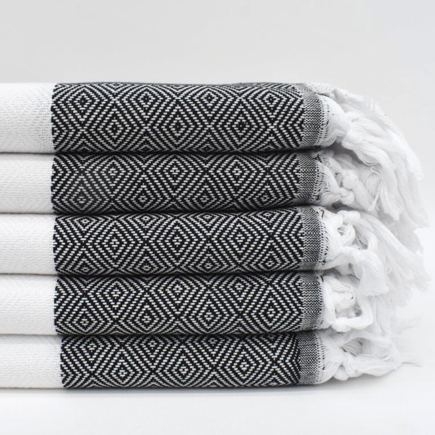 Stack of folded black Sultan Turkish Towels with intricate geometric pattern and white tassels, ideal for luxurious comfort