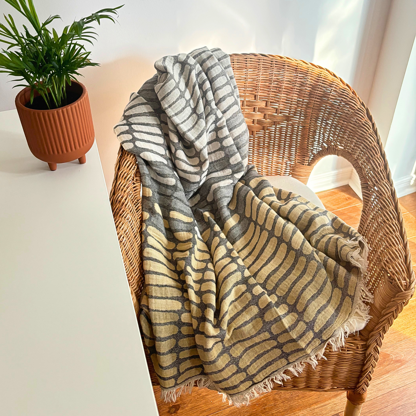 Beige WAVES Turkish Towel draped over a wicker chair next to a potted plant on a wooden floor. The towel features a unique wave pattern with beige and grey colours